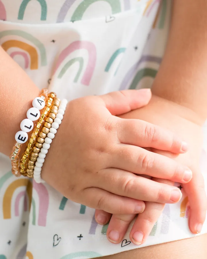 Small hands crossed over a rainbow-print dress, wearing a white letter bead bracelet spelling Ella, stacked with gold and white seed-bead bracelets